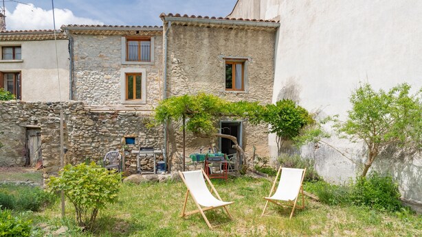 Un bâtiment en pierre avec deux fenêtres et une porte, un petit jardin avec de l’herbe et un arbre, et deux fauteuils de jardin blancs.