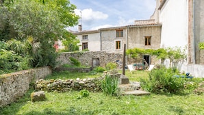 Une cour rustique aux murs de pierre, un petit jardin et un bâtiment traditionnel doté de nombreuses fenêtres.