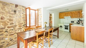Un coin repas avec un mur en pierre, une table en bois et des chaises. On aperçoit, au fond, une cuisine avec des meubles et des appareils électroménagers en bois.