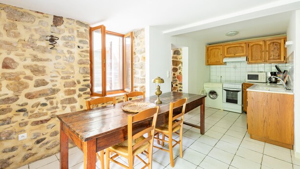 Uma área de refeições com parede de pedra, mesa e cadeiras de madeira. Uma cozinha com armários e eletrodomésticos de madeira é visível ao fundo.