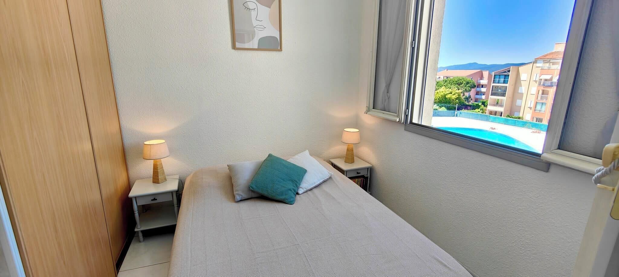 A hotel room with a bed, two bedside tables with lamps, a window with a view of buildings and a pool.