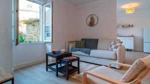 Una sala de estar moderna con un sofá, una mesa de centro y una silla. Un ventanal ofrece luz natural, y se distingue una zona de cocina al fondo.