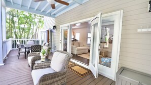 Een overdekt terras met rieten meubilair, een plafondventilator en uitzicht op het interieur door glazen deuren.