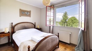 Un dormitorio con cama de madera, mesita de noche con lámpara, ventana con vista a árboles, y un aplique de pared.