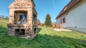 Un horno de ladrillo al aire libre con humo, una casa blanca con contraventanas rojas y un patio cubierto de césped.