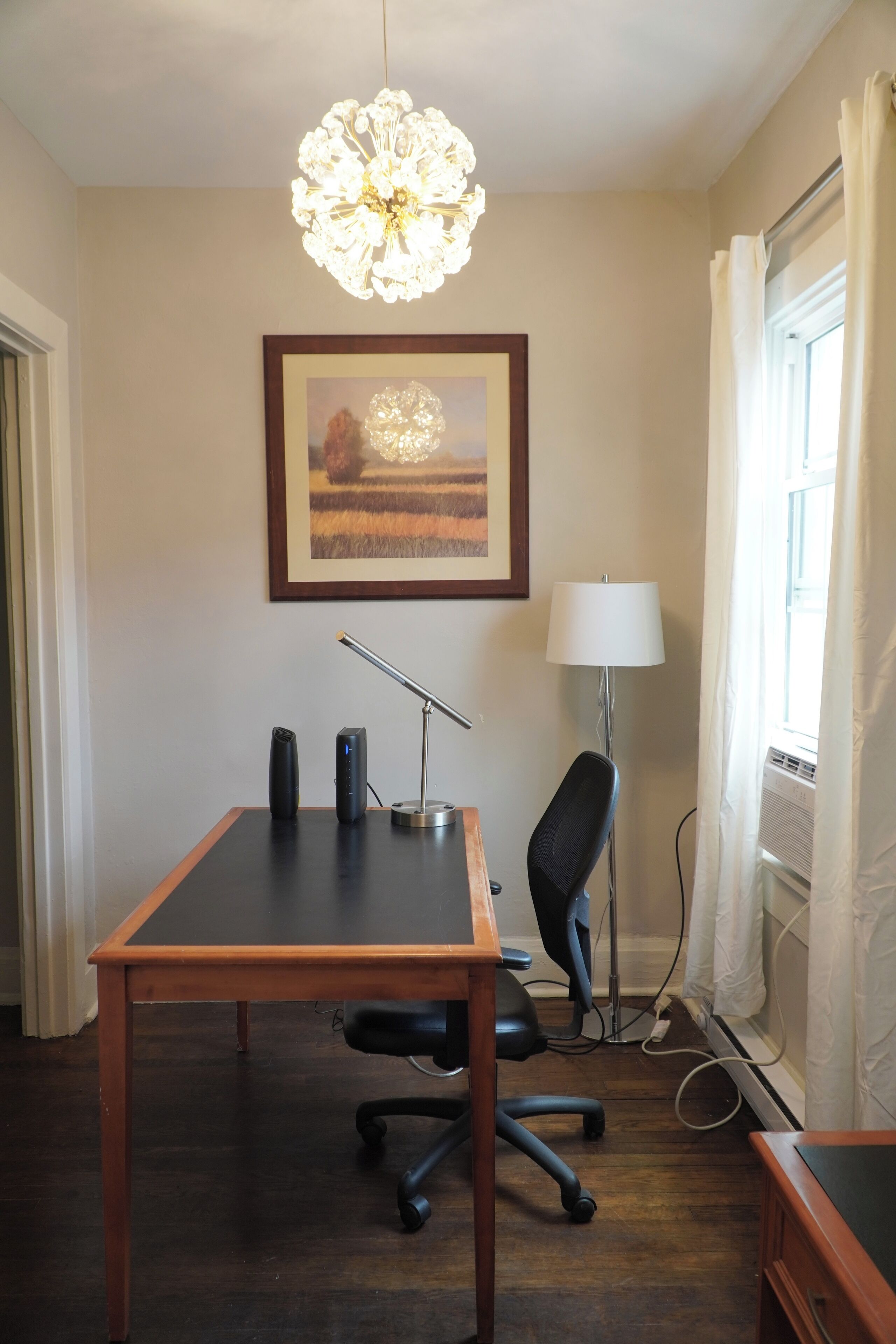 A room with a wooden desk, a black office chair, a standing lamp, a framed picture on the wall, and a window with curtains.
