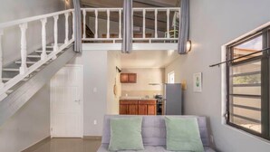Una sala de estar con un sofá gris, una escalera, una cocina y un ventanal con persianas.