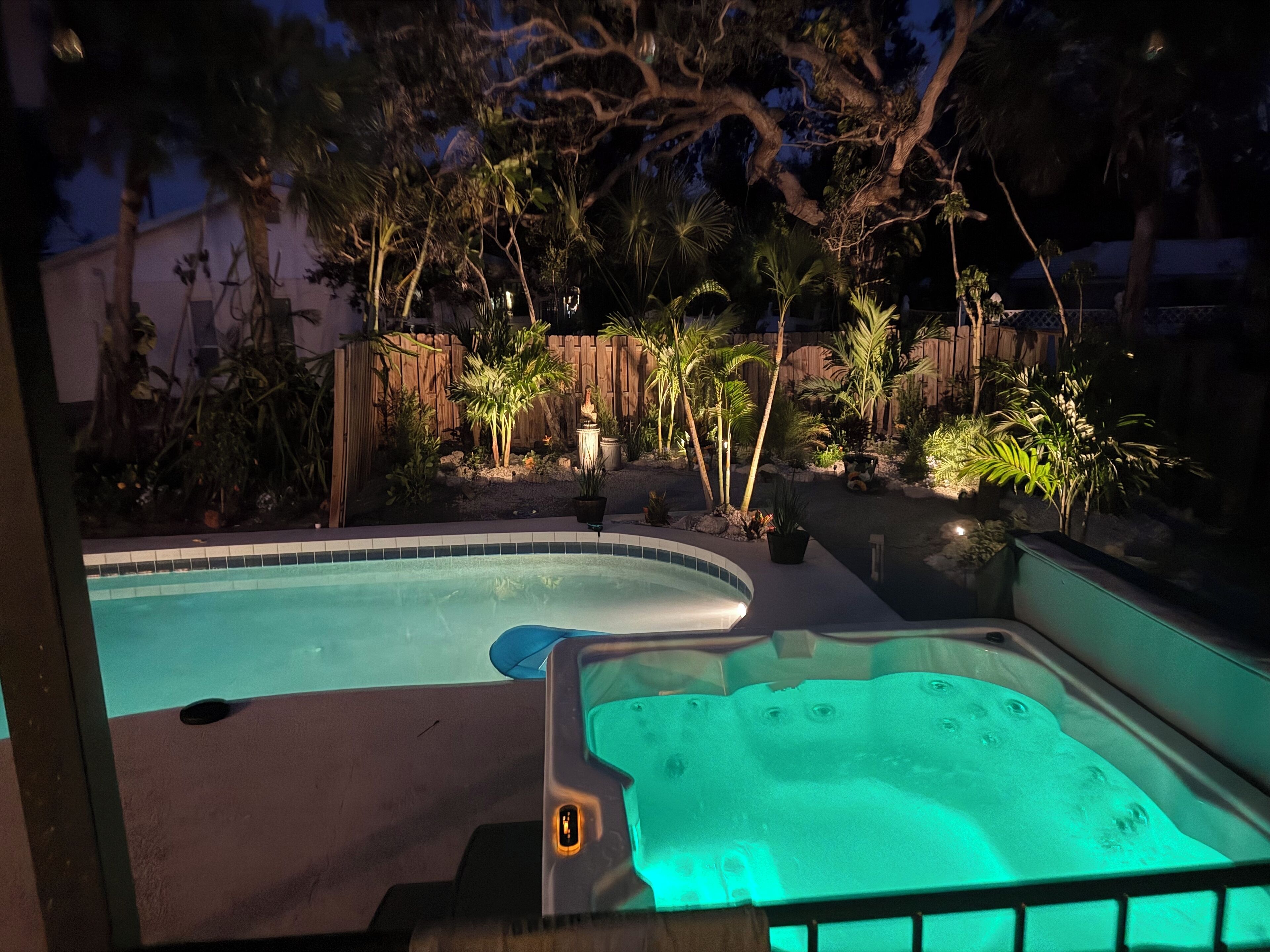 A swimming pool with a hot tub, surrounded by tropical plants and a wooden fence.