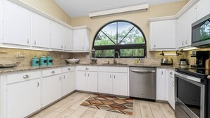A modern kitchen with white cabinets, a stainless steel dishwasher, a black stove, and a large window with a view of greenery. - Island Oasis on Manasota Key- Steps to both Gulf & Bay Access, Heated Pool (Englewood)