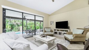 A spacious living room with a white sofa, a coffee table, and wicker furniture. A large TV is mounted on the wall, and there is a view of a swimming pool outside. - Island Oasis on Manasota Key- Steps to both Gulf & Bay Access, Heated Pool (Englewood)
