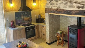 A rustic kitchen with wooden cabinets, a stone wall, a red wood-burning stove, and a dining table with a fruit bowl. - 16th century thatched house, relaxation area for adults and children (Saint-Augustin)