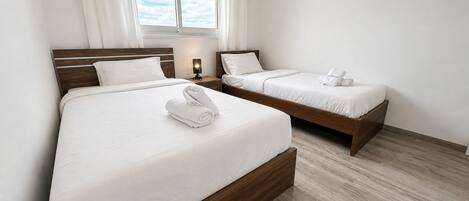 A hotel room with two beds, a window with curtains, a bedside table with a lamp, and an air conditioning unit.