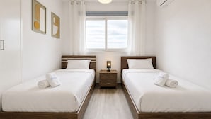 Una habitació d'hotel amb dos llits, una finestra amb cortines, una taula de nit amb una làmpada i un aparell d'aire condicionat.