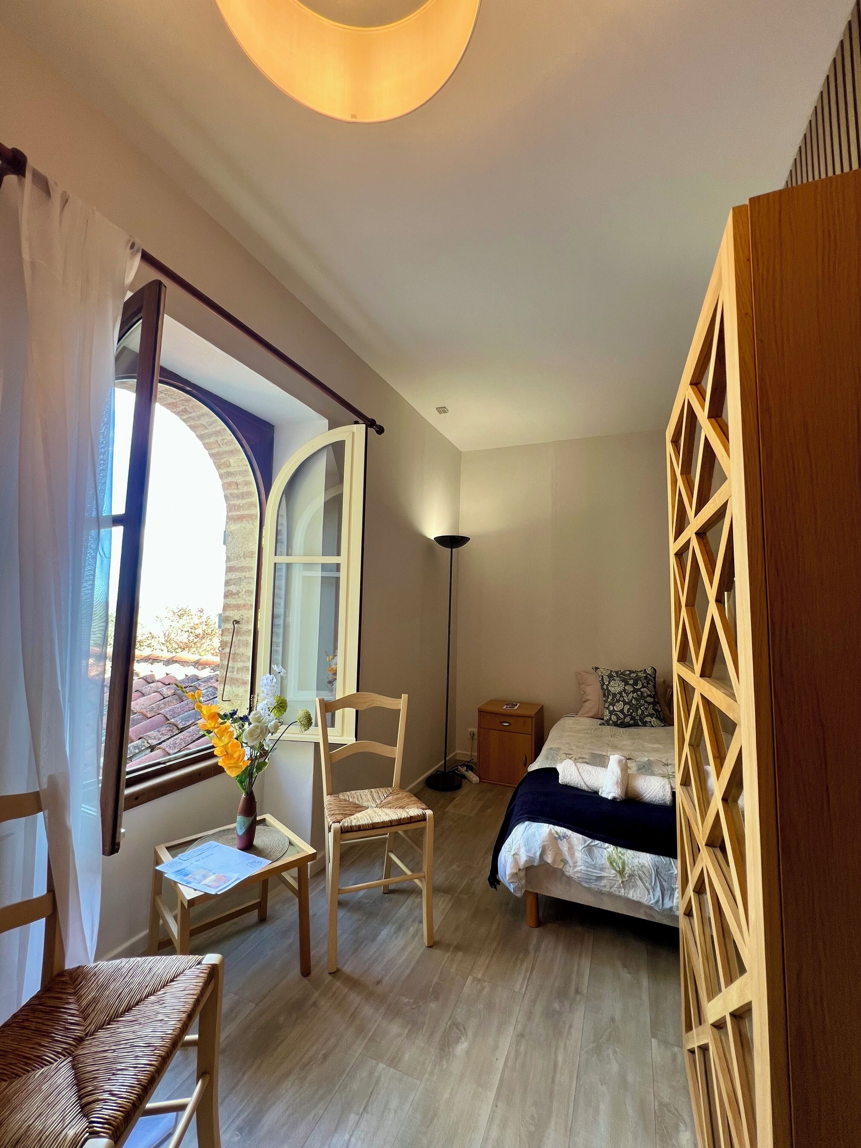 Un dormitorio con cama, una silla, una mesita con un florero y una ventana con cortinas.