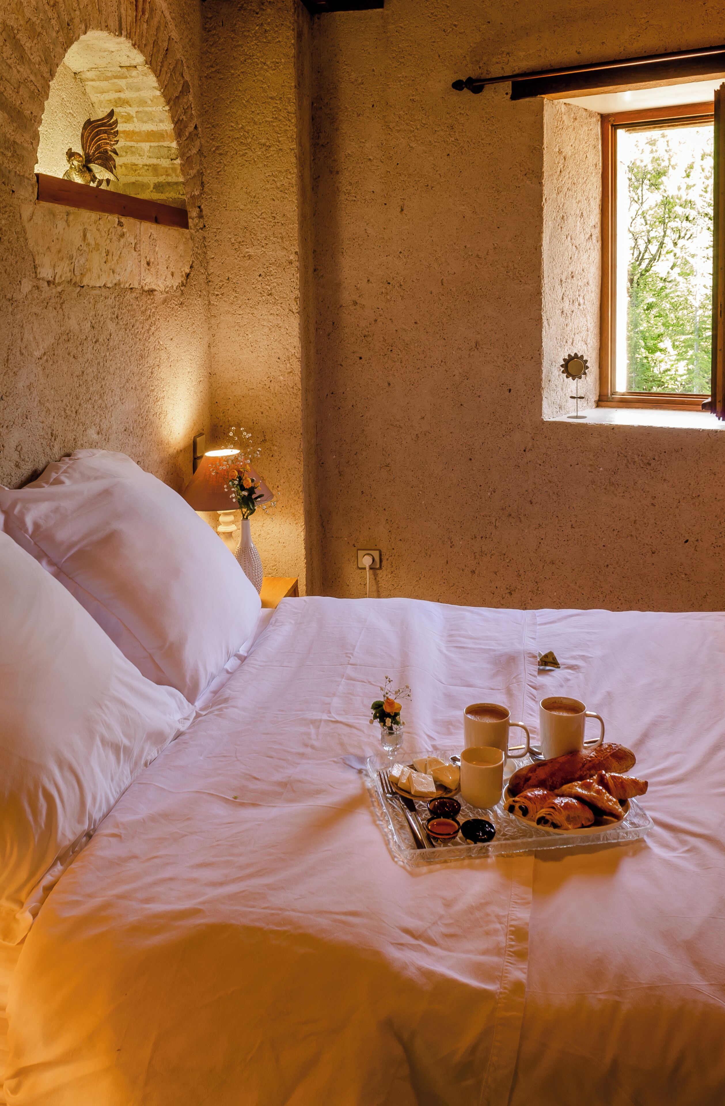 A bed with white linens, a tray of breakfast items, and a window with a view of greenery.