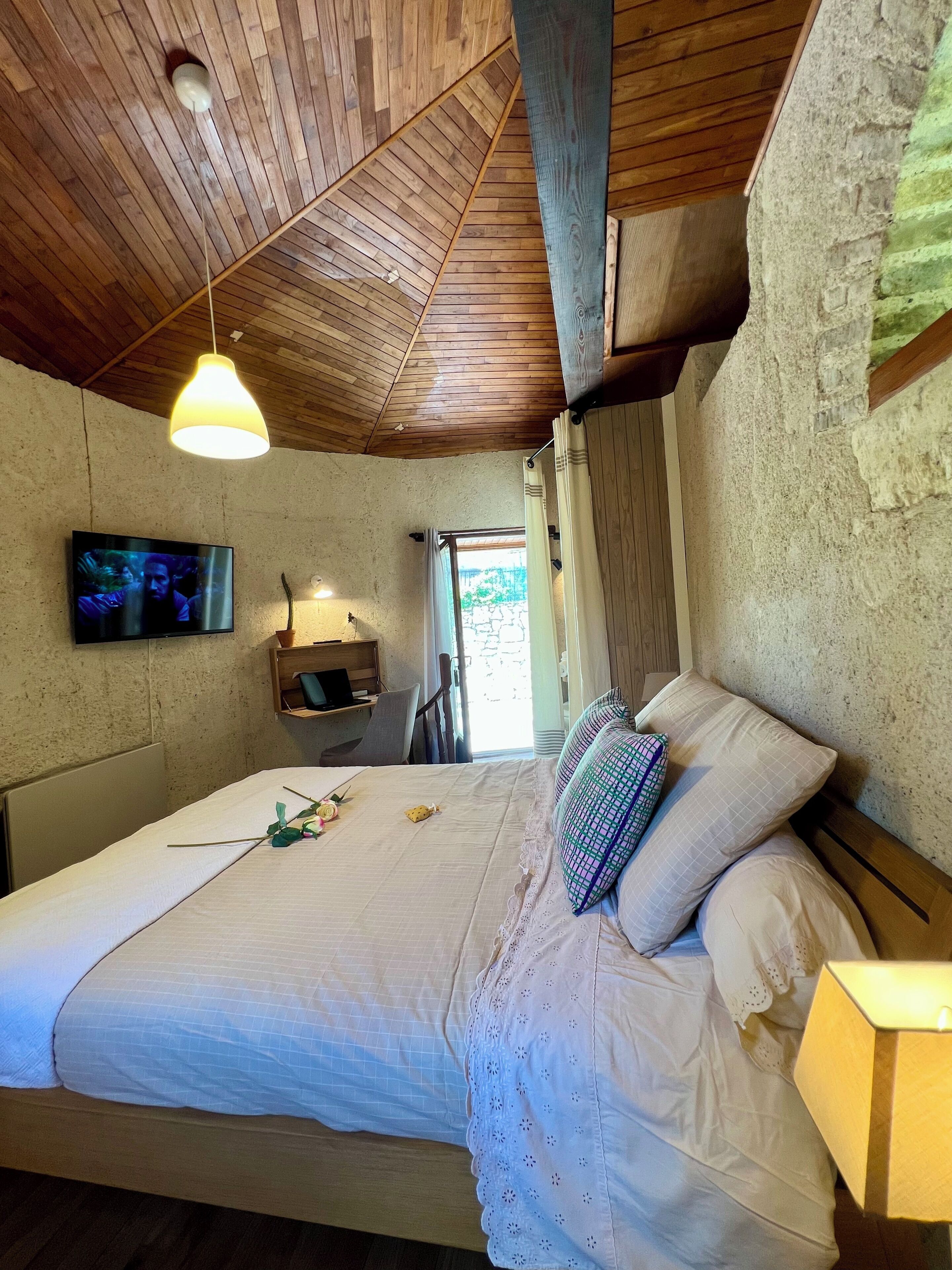 A bedroom with a wooden ceiling, a bed with white linens, a television on the wall, and a window with curtains.