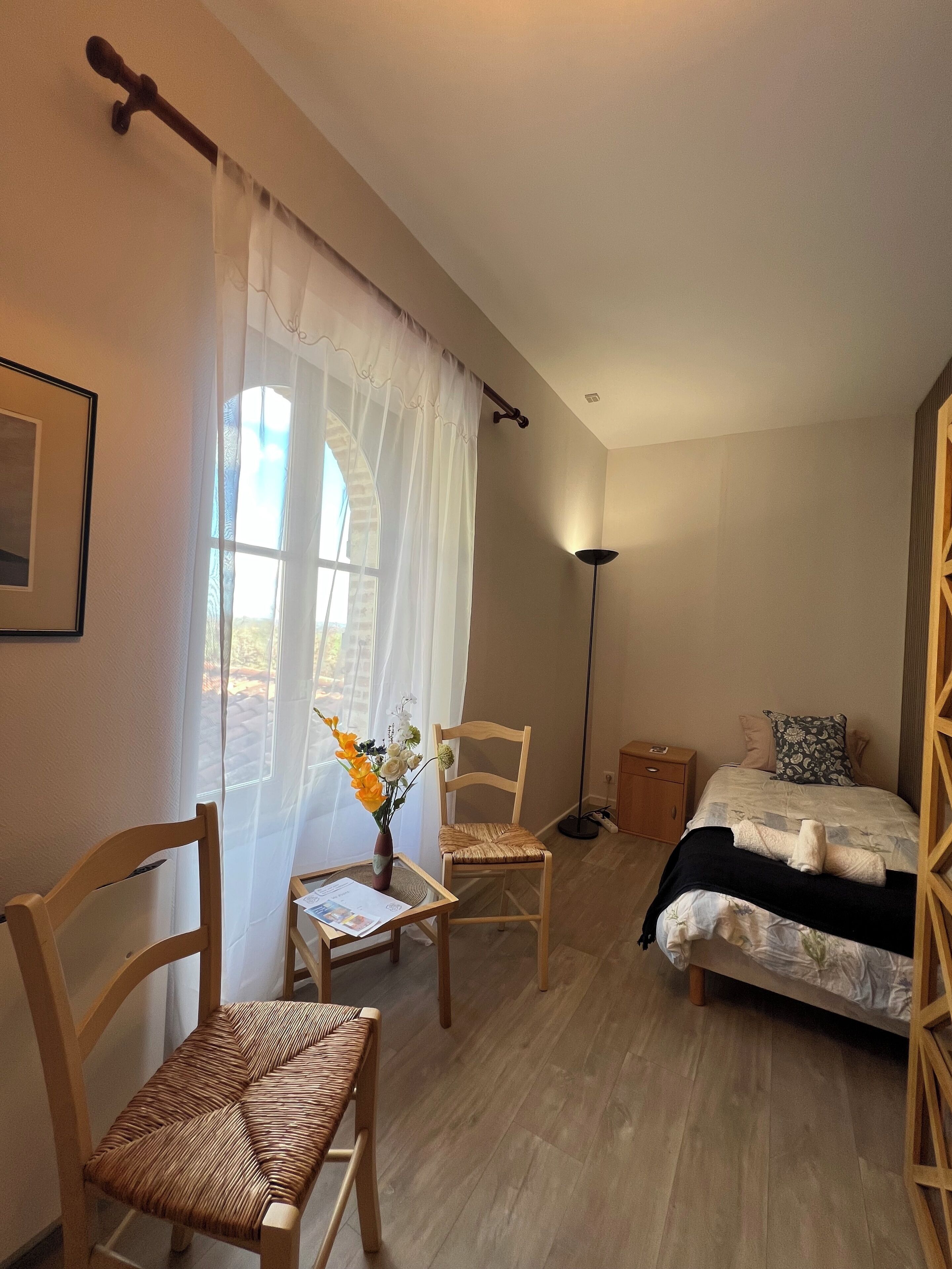 A bedroom with a bed, two wooden chairs, a small table with a vase of flowers, and a floor lamp.