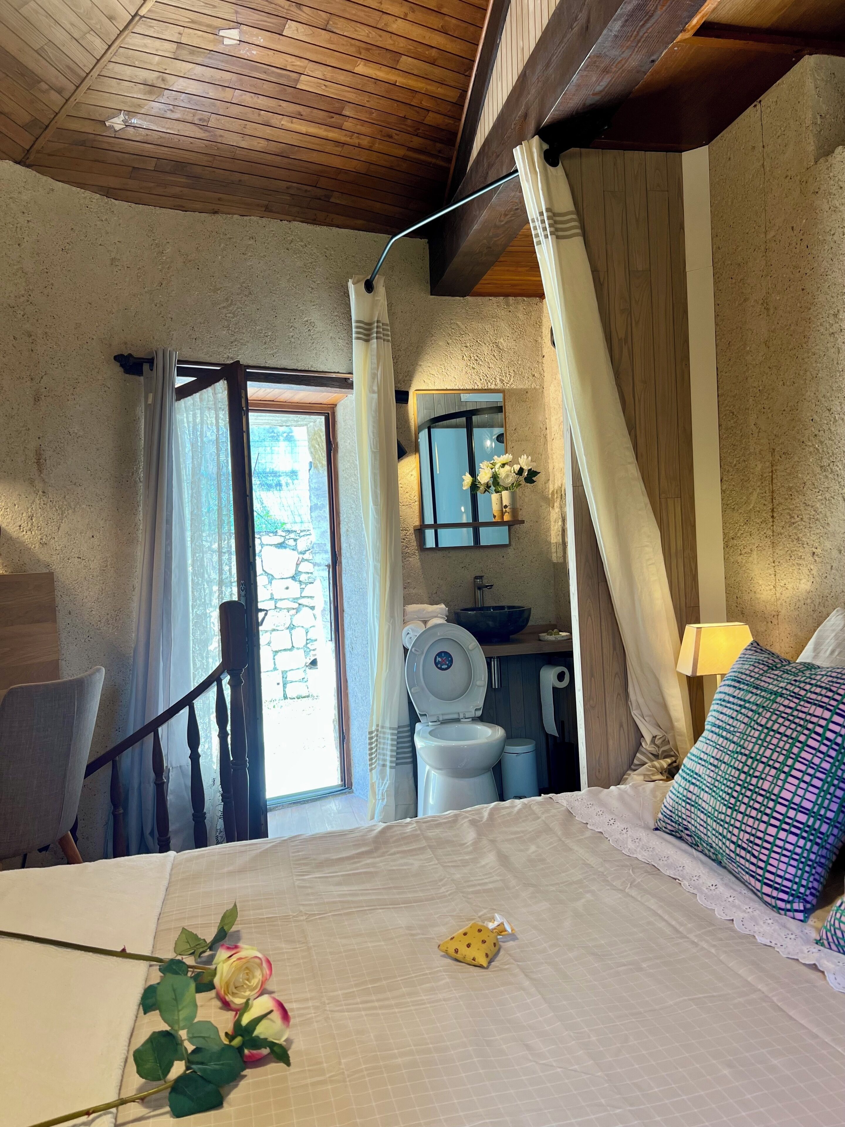 A bedroom with a wooden ceiling, a bed with a white bedspread, a bedside table, a mirror with a vase of flowers, a bathroom with a toilet and sink, and a window with curtains.