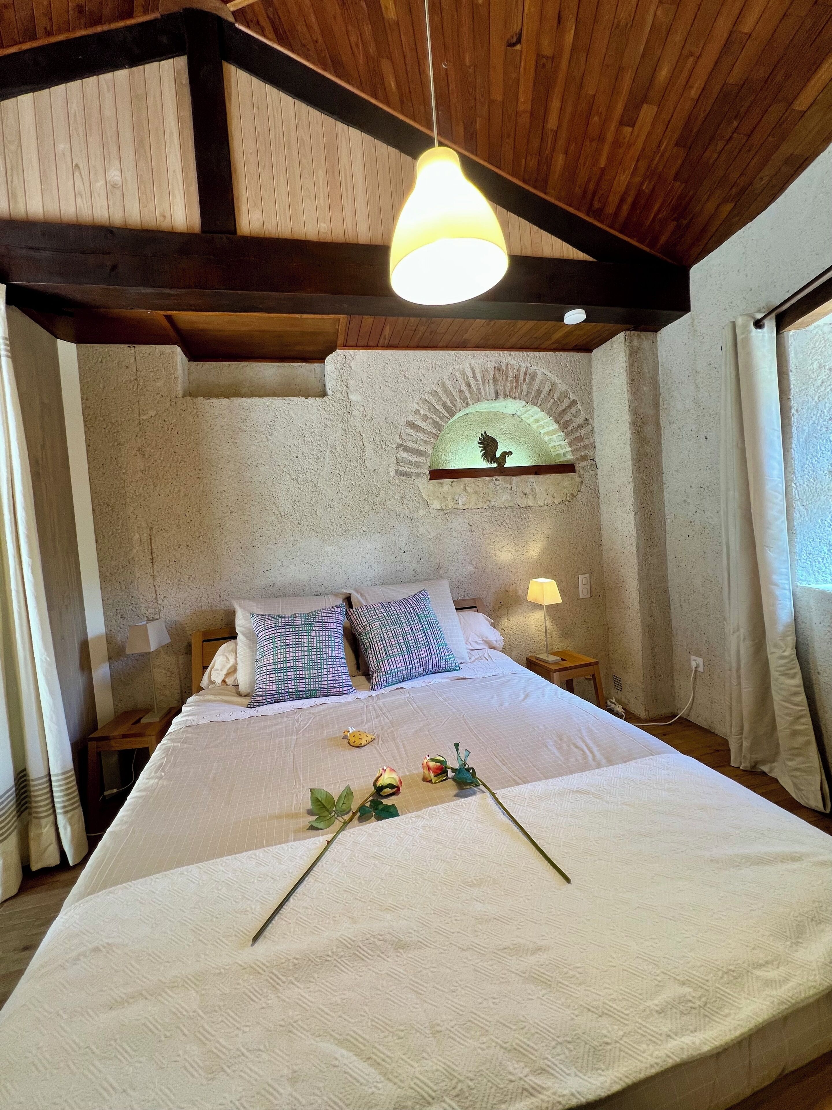 A bedroom with a large bed, a wooden ceiling, a stone wall, and a small table with a lamp.