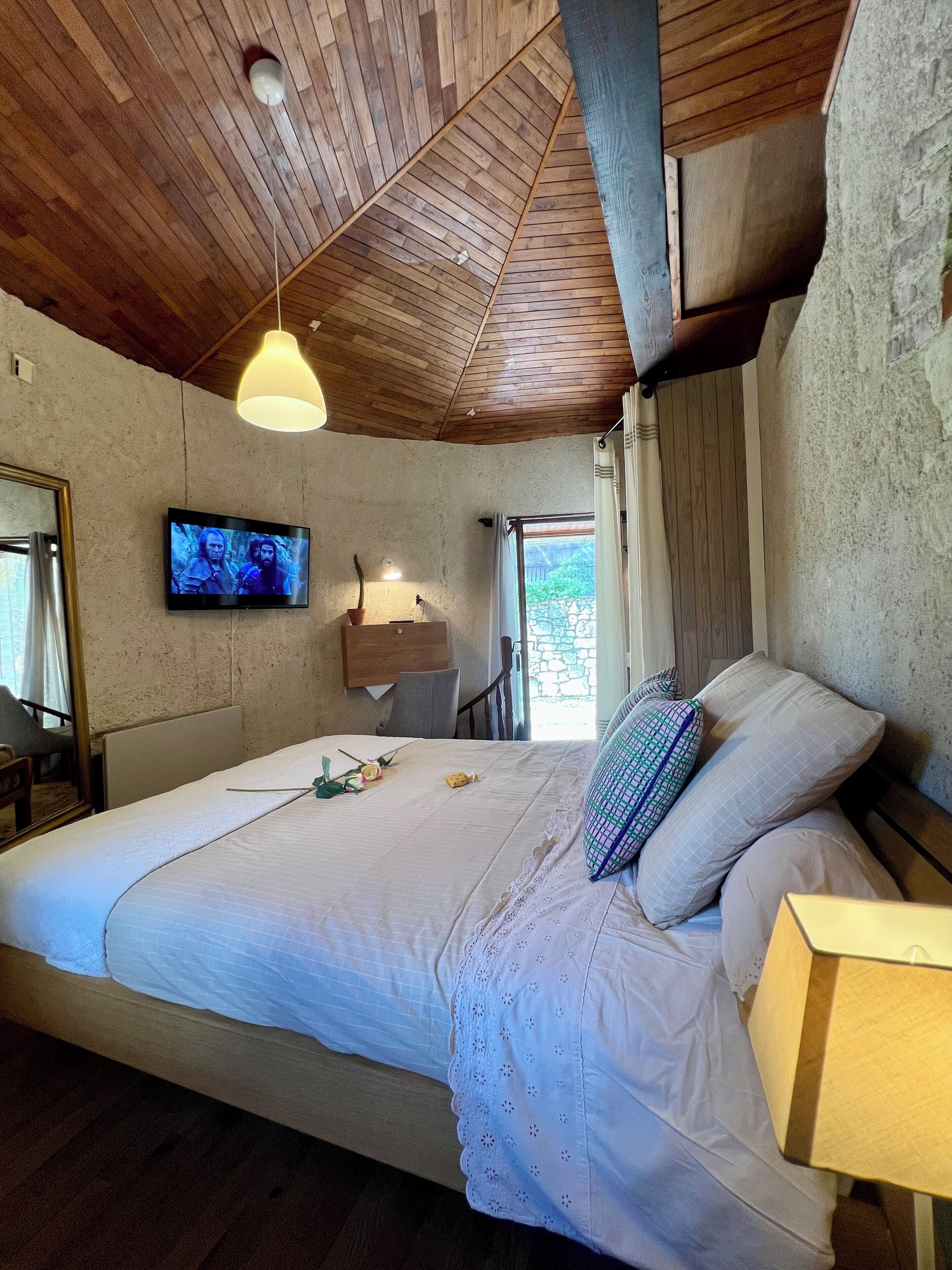 Un dormitorio con techo de madera, cama con ropa de cama blanca, televisor en la pared y ventana con cortinas.