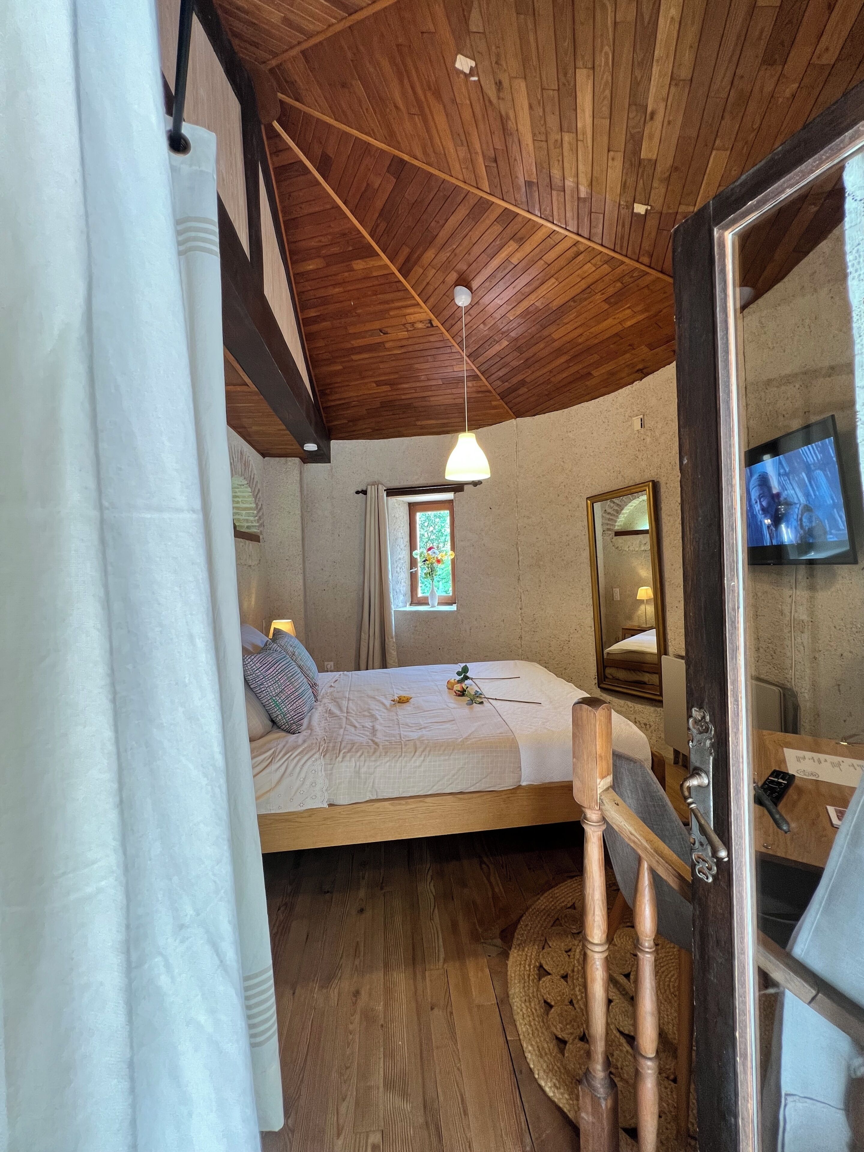 A bedroom with a wooden ceiling, a bed with pillows, a bedside table, a mirror, a window with curtains, and a television on the wall.