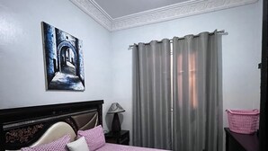 Een slaapkamer met een bed, hoofdbord, nachtkastje, lamp, een aan de muur hangend schilderij, een raam met gordijnen en een kamer met een patroon.