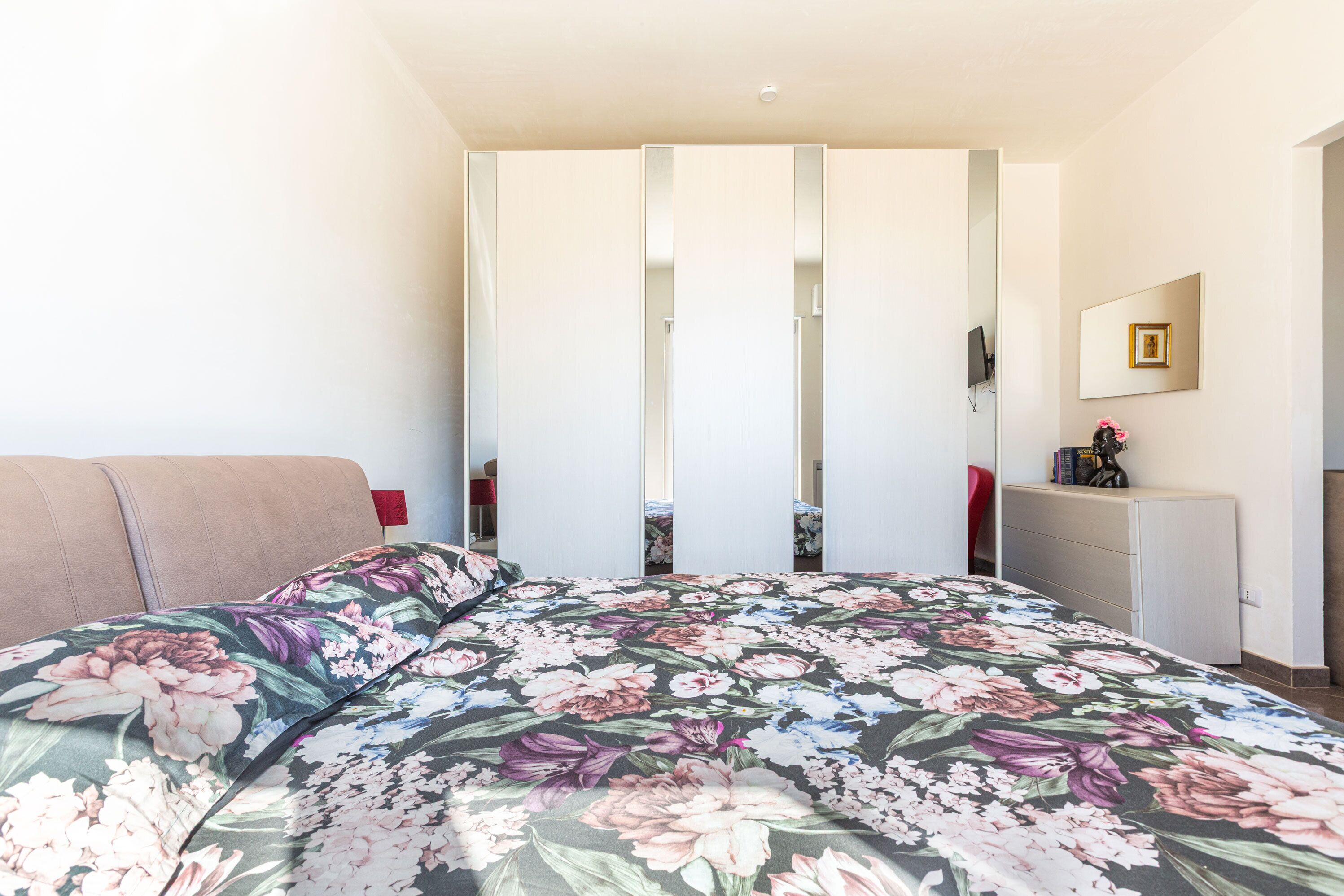 Un dormitorio con colcha estampada, un armario blanco, un escritorio con espejo y una silla roja.