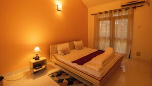 A bedroom with a bed, a nightstand with a lamp, a window with curtains, and an air conditioning unit. - 5 Min to Benaulim Beach SERENE ESCAPE VILLA (Benaulim)