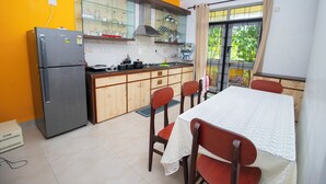 A compact kitchen with a refrigerator, wooden cabinets, a gas stove, and a dining table with chairs. - 5 Min to Benaulim Beach SERENE ESCAPE VILLA (Benaulim)