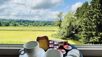 Un plateau avec deux tasses et une assiette de nourriture, comprenant du beurre et de la confiture, est posé sur une table près d’une fenêtre donnant sur un champ verdoyant et des arbres.