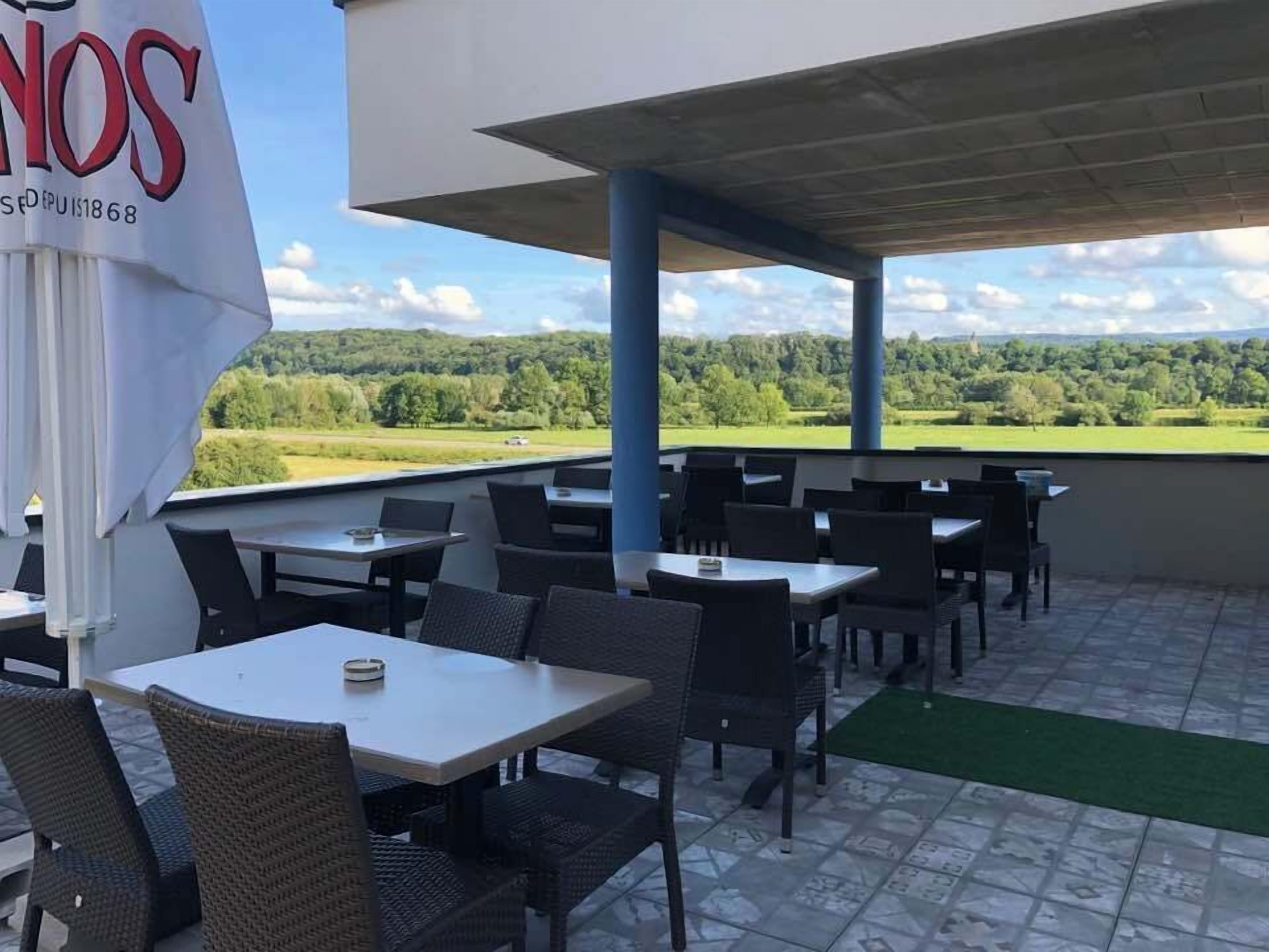 Een terras buiten met tafels en stoelen, uitzicht op een groen landschap, en een bord met de tekst 'NOS DEPUIS 1868'.