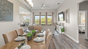 A dining area with a wooden table set for four, surrounded by beige upholstered chairs. There is a ceiling fan above and a large window with curtains. - Movie Theater/Game Room, FREE Pool Heat! No Rear Neighbors, Minutes to Disney! (Kissimmee)