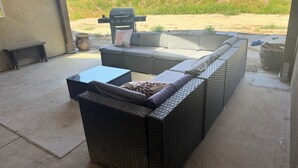 Une terrasse couverte, meublée avec des meubles en osier, dotée d’un barbecue et offrant une vue sur une zone verdoyante.