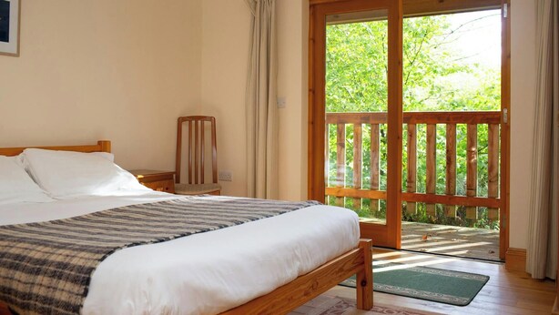 A bedroom with a wooden bed, a chair, a nightstand, a window with curtains, and a view of greenery outside. - Robin's Perch Eco Lodge Apartment, Lampeter (Lampeter)