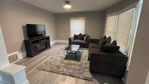 A living room with a flat-screen TV, dark sofas, a coffee table, and a patterned rug. - Just remodeled, for you! (Grand Rapids)