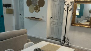 Un coin repas avec une table blanche, des chaises et un élément décoratif central. On y trouve un porte-manteau avec un miroir, une porte dotée d’une poignée décorative et une étagère avec des chapeaux.