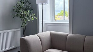 Un salon avec un canapé beige, un lampadaire, une plante en pot et une fenêtre donnant sur le ciel et les arbres.