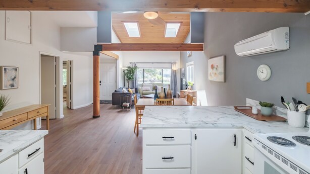 Una cocina moderna con armarios blancos, una encimera de mármol y una estufa integrada. Hay un comedor con mobiliario de madera y un ventanal amplio.