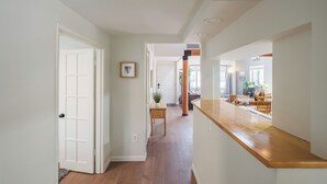 Un pasillo con suelo de madera, paredes blancas y un mostrador de madera a la derecha. En el lado izquierdo hay una puerta y una pequeña mesa con una planta en el centro.