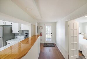 Cozinha moderna com armários brancos e bancada de madeira, um corredor que leva a um quarto com porta branca e mesa de cabeceira.