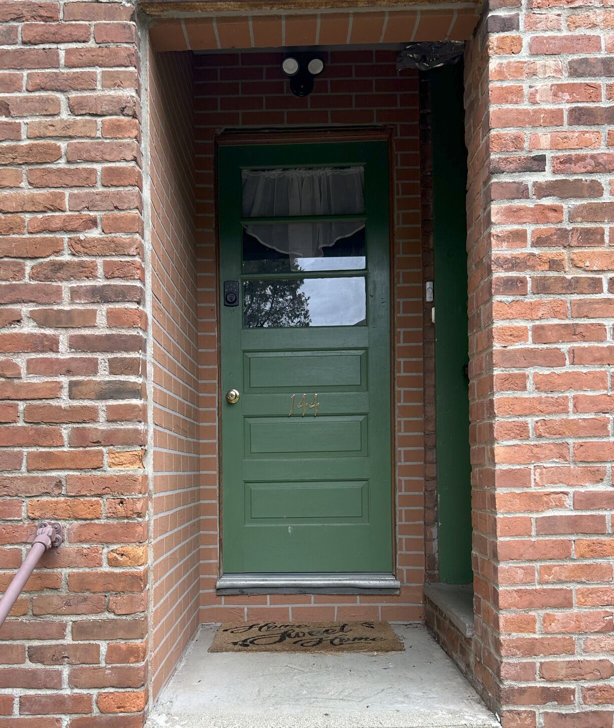 Une porte verte avec une plaque portant un numéro, une petite fenêtre avec un rideau et un paillasson sur lequel est écrit : « Chez soi, on est bien ».