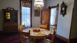 Une salle à manger avec une table en bois, une nappe à motifs floraux et des chaises. On y trouve un lustre, un buffet en bois et une horloge au mur.