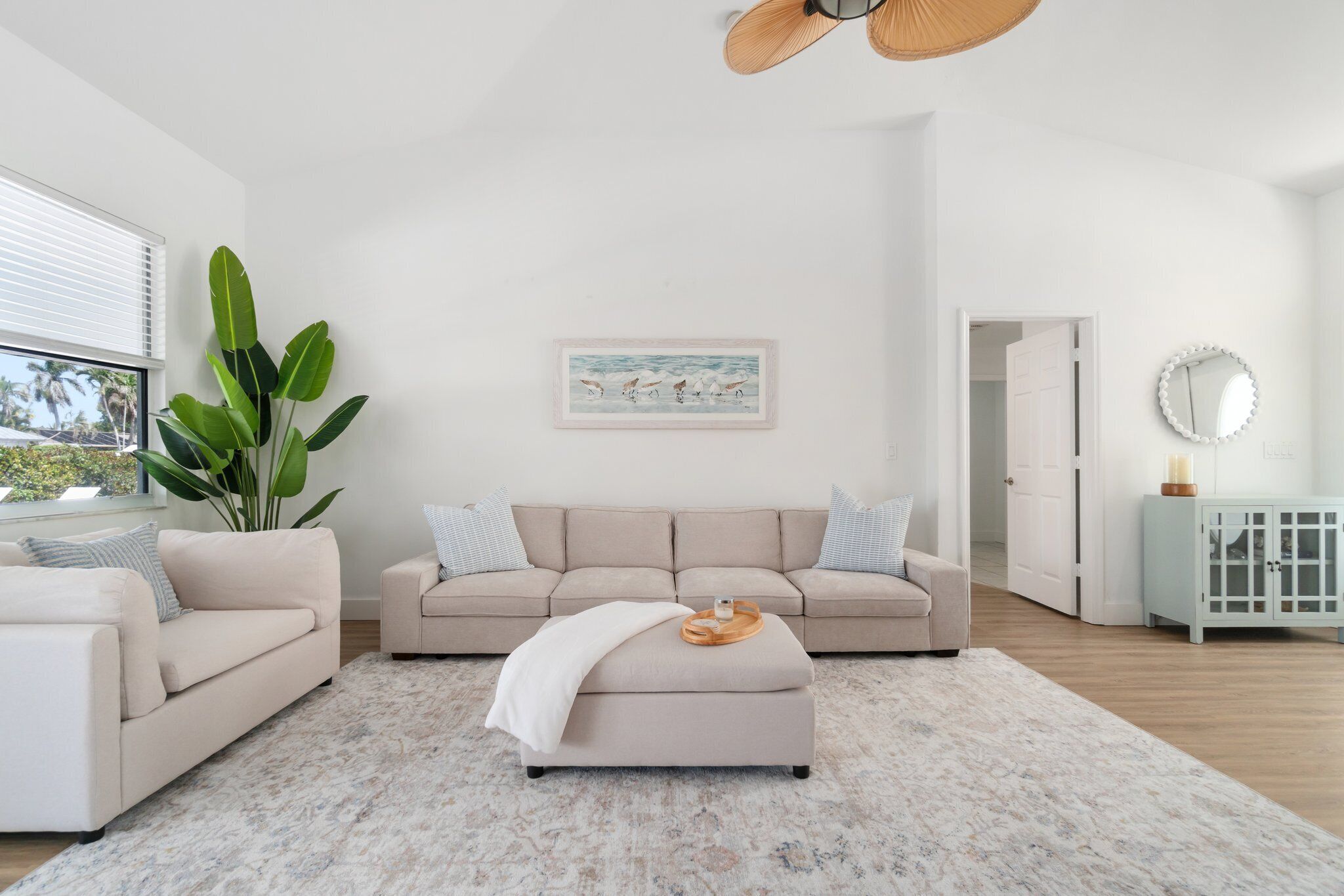 A modern living room with a beige sofa, a matching ottoman, and a large potted plant.
