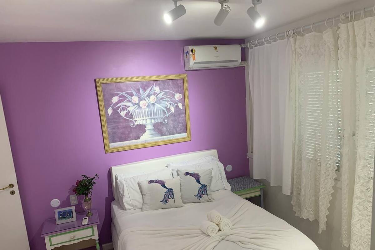 Quarto com parede roxa, cama branca com almofadas decorativas, mesa de cabeceira com uma planta e ventilador de teto.