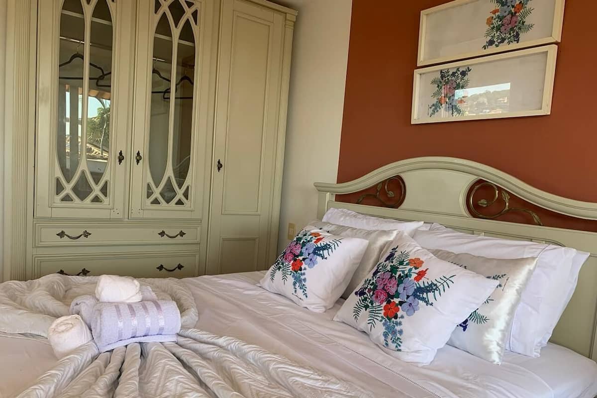 Uma cama bem arrumada com almofadas florais, cabeceira bege e um guarda-roupa grande com portas de vidro.