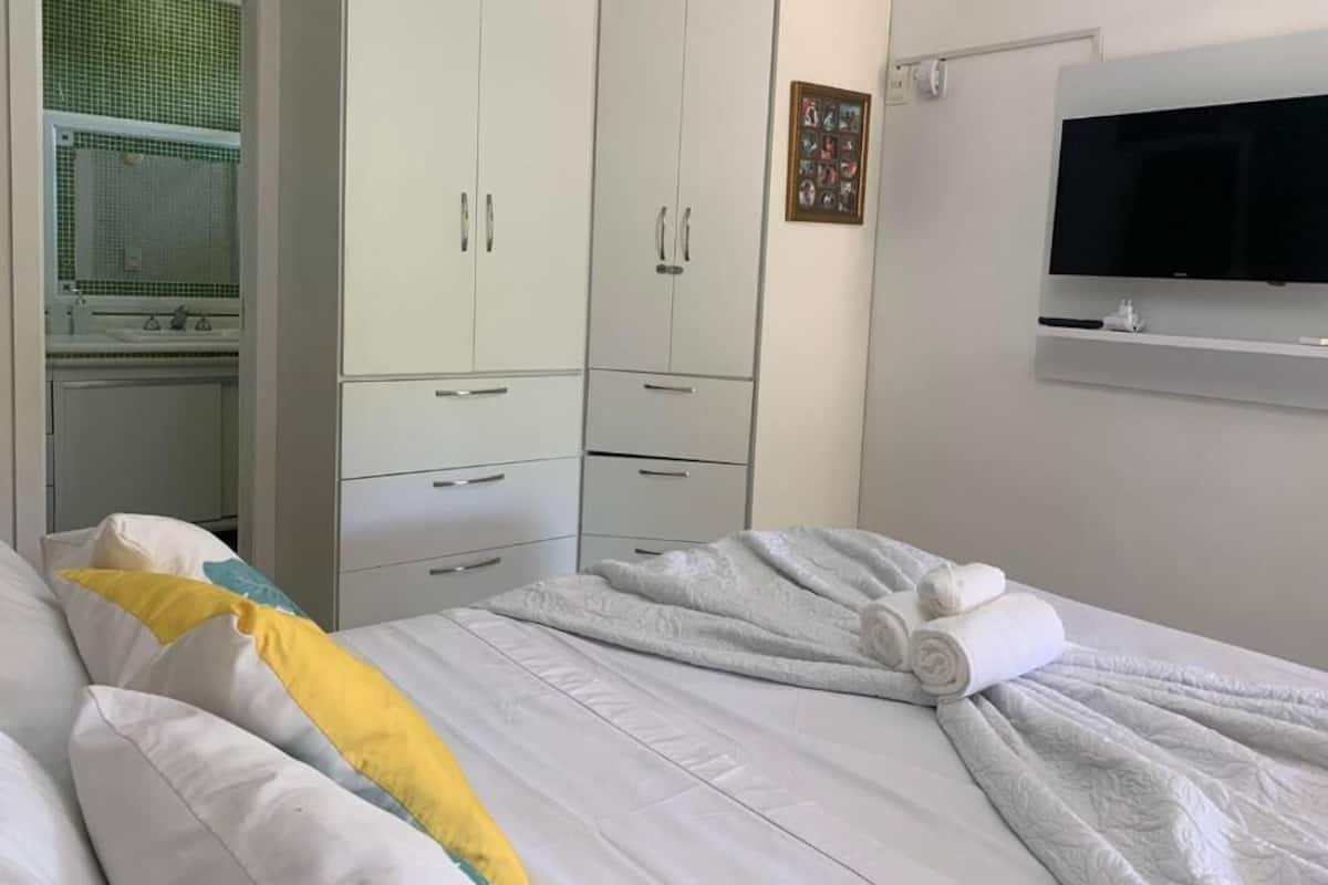 Quarto de hotel com cama, guarda-roupa branco e televisão na parede.