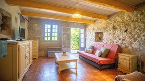 A cozy room with a stone wall, a wooden ceiling, a sofa, a table, and a TV on a cabinet.