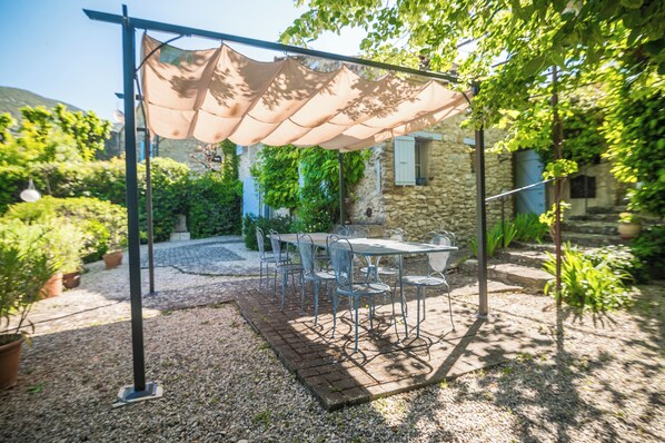 A covered outdoor seating area with a table and chairs, surrounded by potted plants and a stone building in the background. - Le Refuge Provençal Nijonyas (Malaucène)