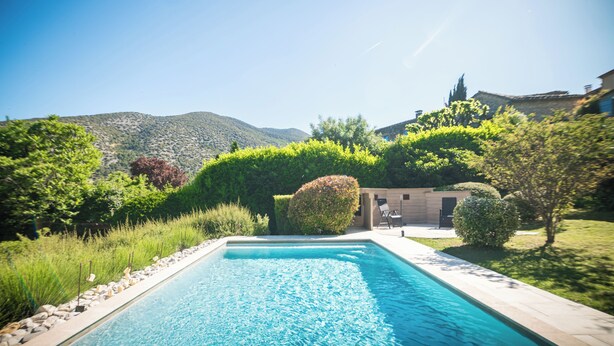 A rectangular swimming pool with a clear blue water surface, surrounded by a stone-lined edge and a grassy area with bushes and trees.