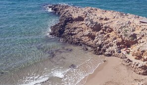 Une côte rocheuse bordée d’une eau claire, d’une plage de sable et d’une petite construction située sur la droite.